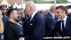 Президент США Джо Байден (справа) тисне руку президенту України Володимиру Зеленському (ліворуч), а президент Франції Еммануель Макрон (праворуч) спостерігає під час відзначення річниці висадки союзників у Нормандії, 6 червня 2024 року 