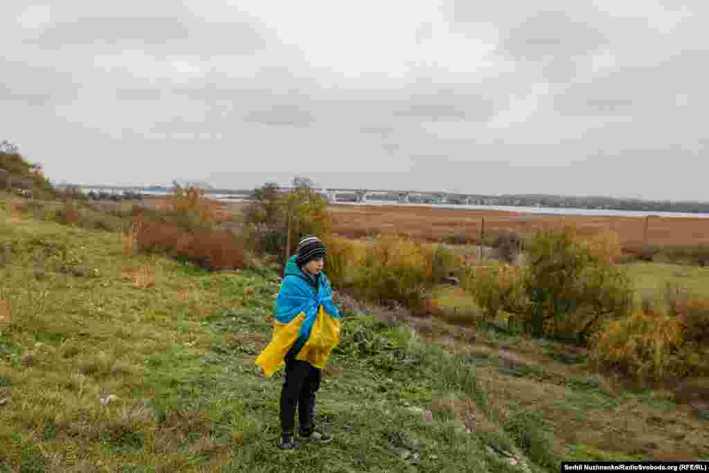 Хлопчык, ахінуты ўкраінскім сьцягам, стаіць непадалёк ад зруйнаванага Антонаўскага моста ў першыя дні вызваленьня Хэрсона ад расейскіх войскаў. Лістапад 2022 году