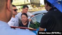 Сотрудник полиции и неизвестный с зонтом наблюдают, как люди в штатском усаживают женщину в полицейский автомобиль во время задержаний на площади Аль-Фараби. Шымкент, 6 июля 2019 года.