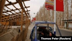 Человек в припаркованной вдоль дороге машине под плакатом с государственным флагом Китая и изображением президента страны Си Цзиньпина. Хотан, 21 марта 2017 года.