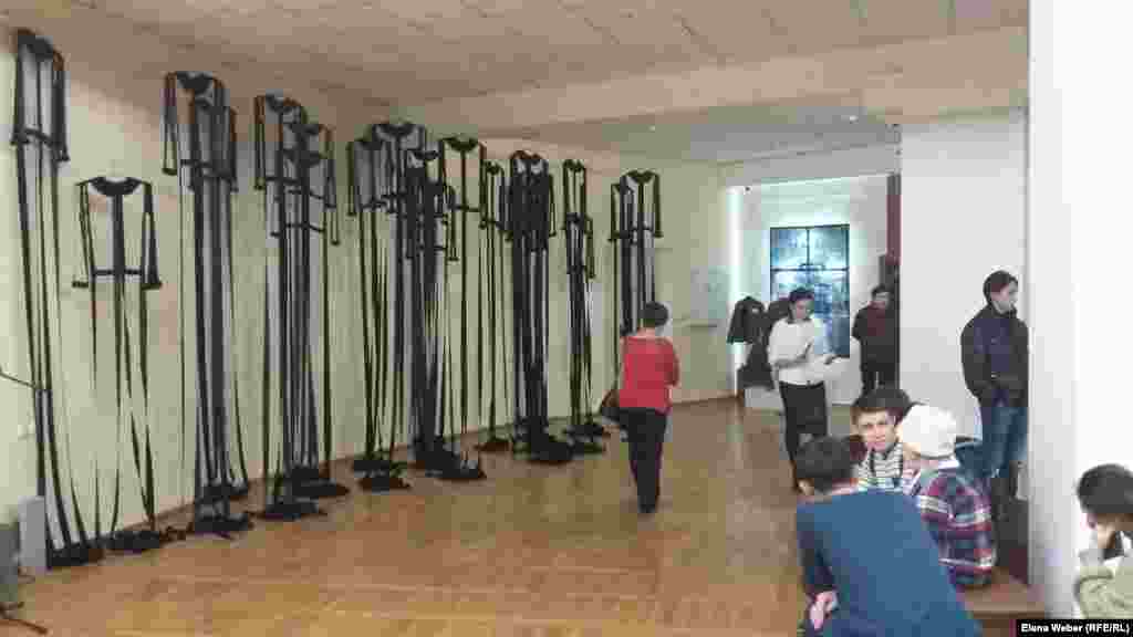 Эта инсталляция &quot;АЛЖИР&quot; посвящена 18 тысячам узниц Акмолинского лагеря жен изменников родины. Не все посетители сразу понимали, что это, но приглядевшись, видели силуэты женских платьев. Образ длинных лагерных черных балахонов-платьев, от которых остались лишь швы, воротнички и манжеты, выбран автором не случайно. По задумке Айнур Саденовой, несмотря на потерю семьи, нечеловеческие условия в лагерях и физическую надломленность, узницы оставались женственными, добрыми и интеллигентными.