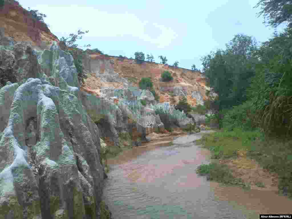 Ручей, окруженный песчаными глыбами из разноцветного песка. На дне ручья видны черные пятна - следы титана, который добывают неподалеку от ручья.