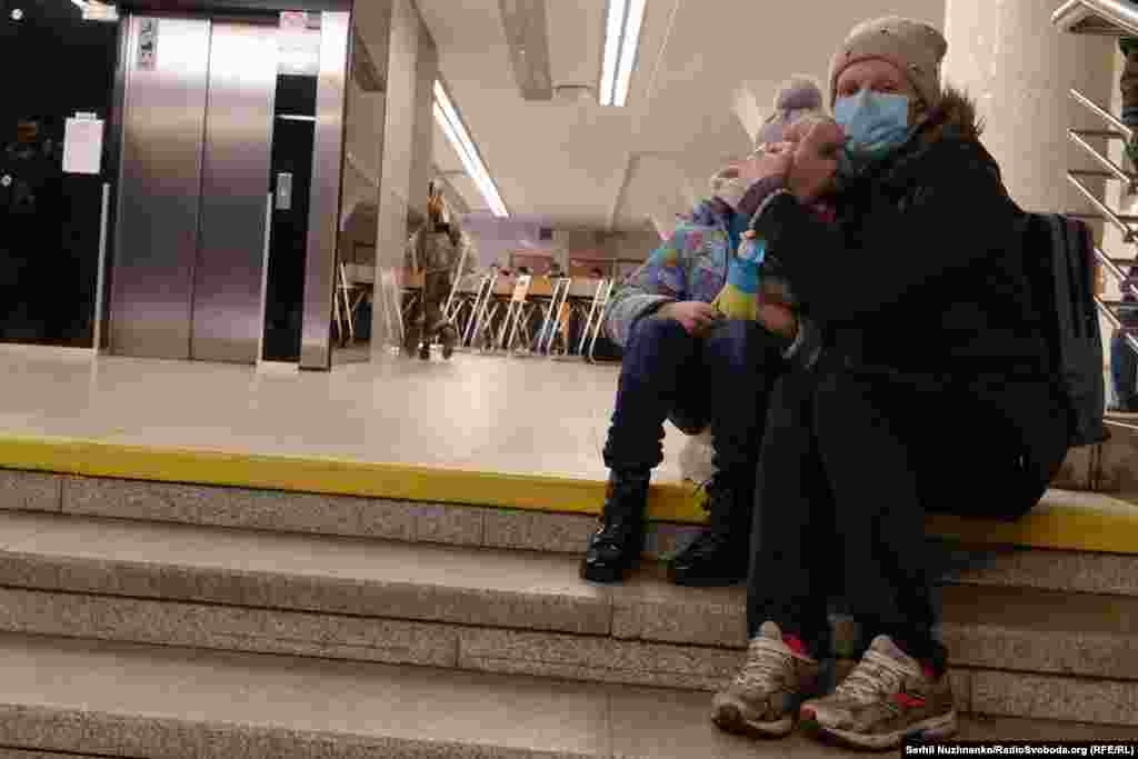 Жанчына абдымае дзіця, хаваючыся ад абстрэлаў на станцыі мэтро «Галасееўская», Кіеў, 24 лютага 2022 году. Першыя гадзіны поўнамаштабнага ўварваньня: адны людзі яшчэ звыкла едуць у мэтро па справах, а іншыя ўжо сьпяшаюцца з валізамі выехаць з гораду