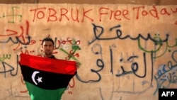 Демонстрант у Тобруку на сході Лівії тримає старий «дореволюційний» прапор країни часів монархії, яку скинув переворот Каддафі. На стіні написи: «Тобрук сьогодні вільний», «Геть Каддафі!»
